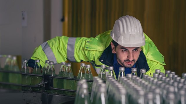 佩戴安全帽的飲料生産廠工人俯身檢查傳送帶上的玻璃瓶